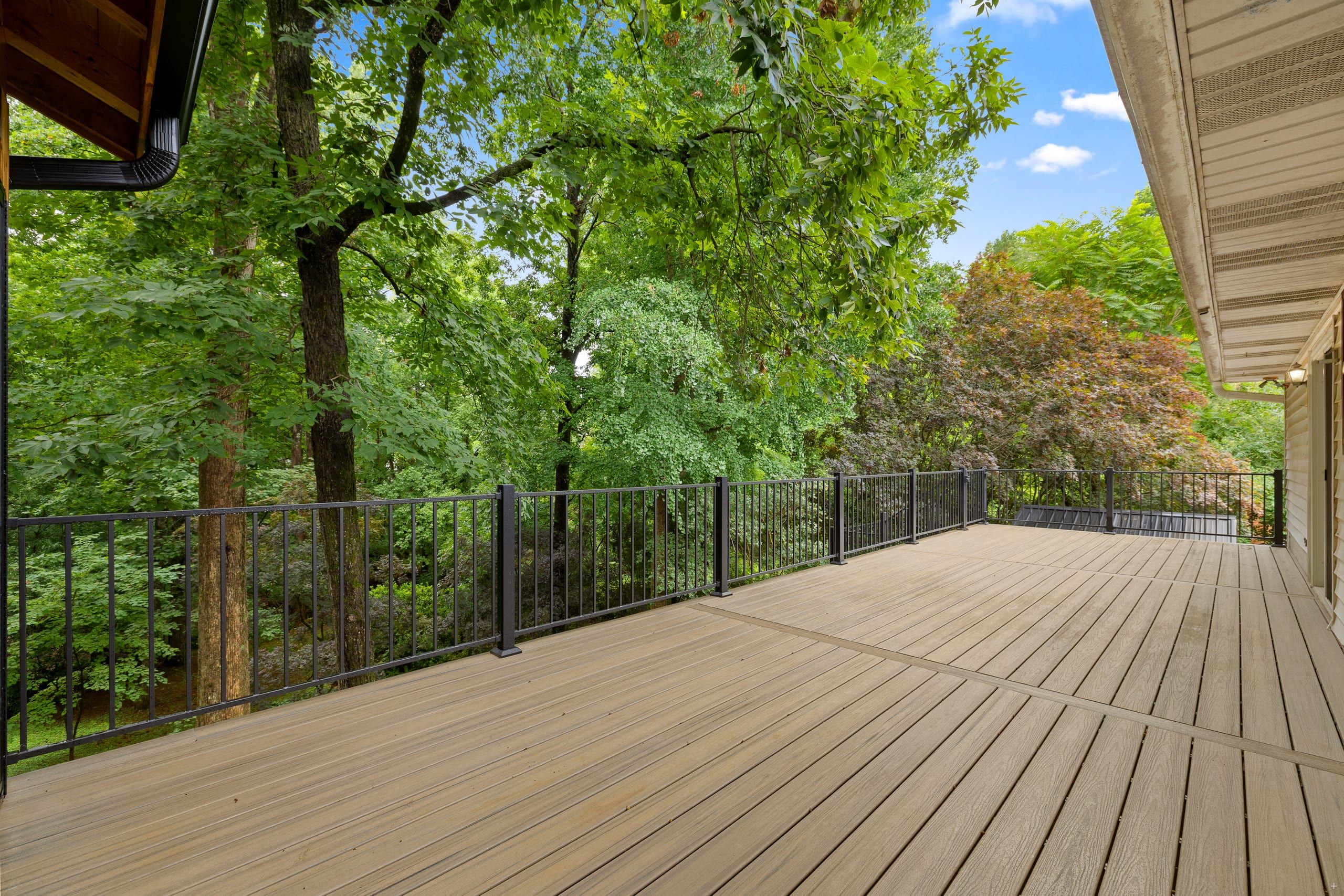 Venice open elevated composite deck with two-tone wood-grain boards, black aluminum railing, and a wooded backyard under blue sky