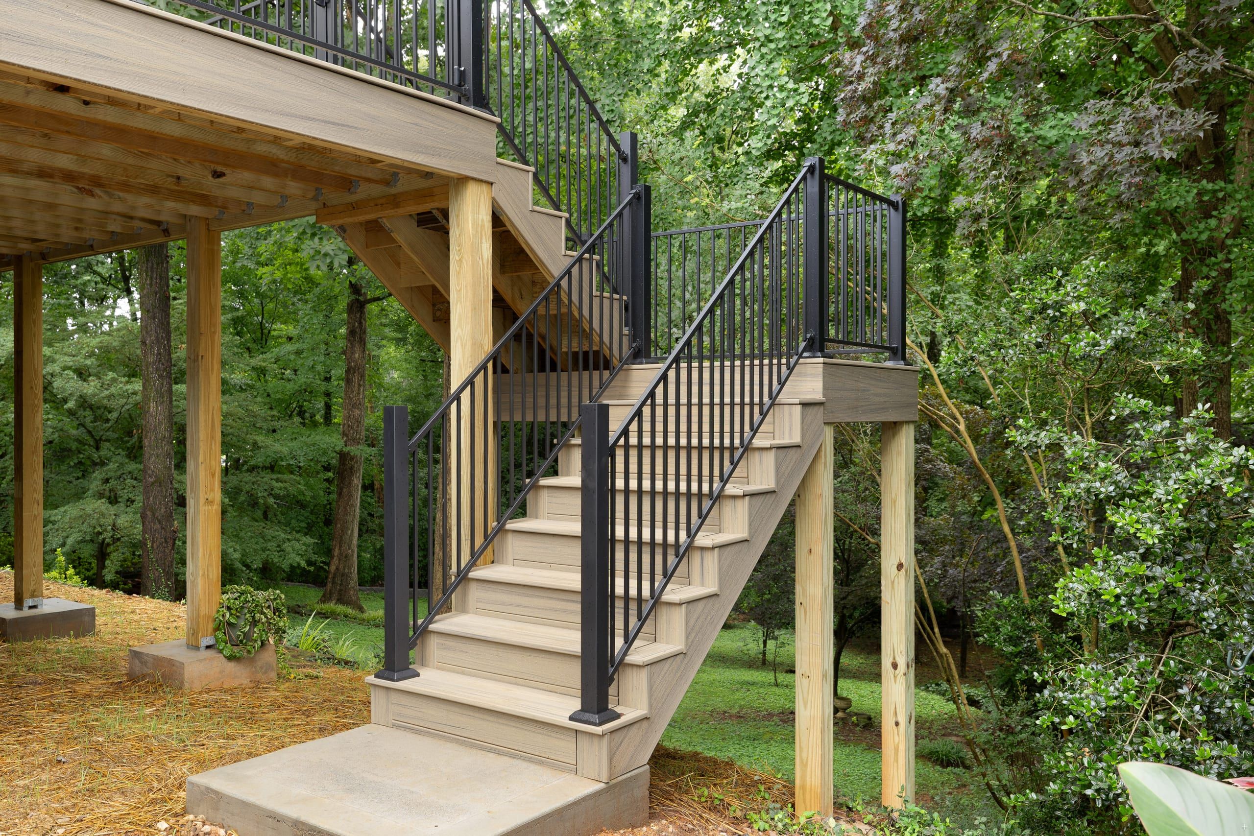 Composite staircase with black aluminum railing descending from the Venice deck to a concrete landing in a wooded yard
